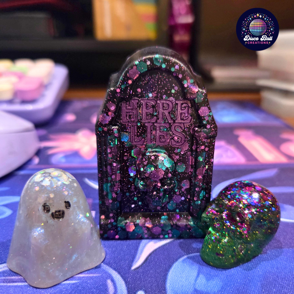Decorative tombstone with 'Here Lies' text, surrounded by glittery ghost and skull figurines on a colourful surface.