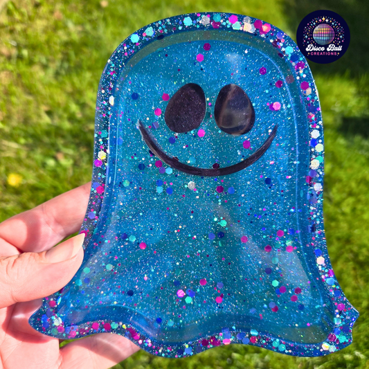 Ghost shaped blue glittery trinket tray with a smiley face held by a hand against a grass background