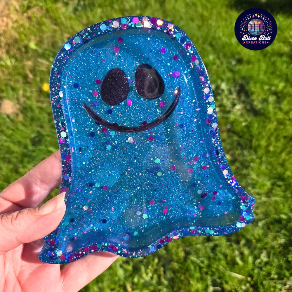 Ghost shaped blue glittery trinket tray with a smiley face held by a hand against a grass background