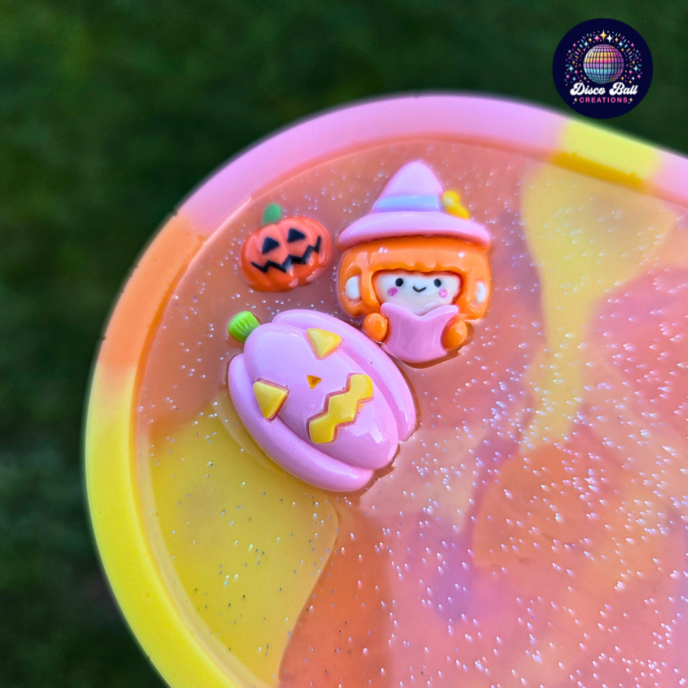 Oval trinket tray made from resin using yellow, pink and orange pastel colours with a subtle glitter in-fill and 3 character charms, a witch, a small pumpkin and a larger pumpkin