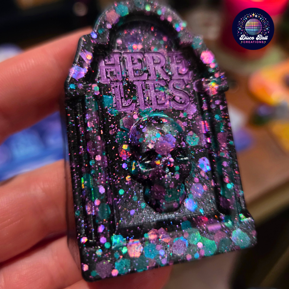 Glittery tombstone-shaped object with 'Here Lies' text held by a hand, brand 'Disco Ball Creations' visible.