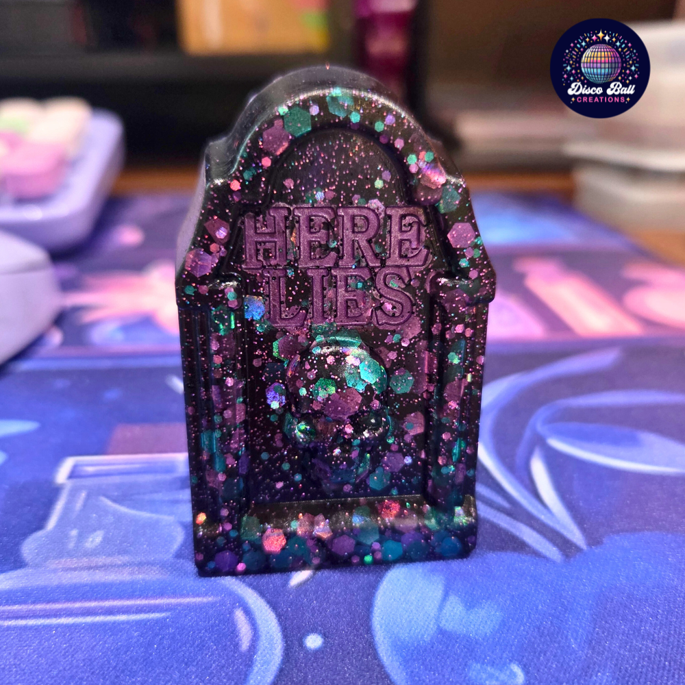 Decorative tombstone with 'Here Lies' text on a colourful surface, featuring glittery purple and blue design.