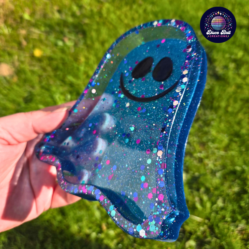 Ghost shaped blue glittery trinket tray with a smiley face held by a hand against a grass background