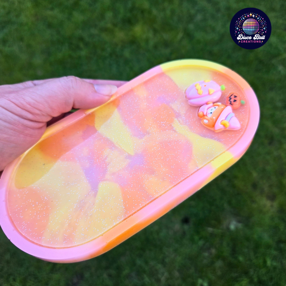 Oval trinket tray made from resin using yellow, pink and orange pastel colours with a subtle glitter in-fill and 3 character charms, a witch, a small pumpkin and a larger pumpkin