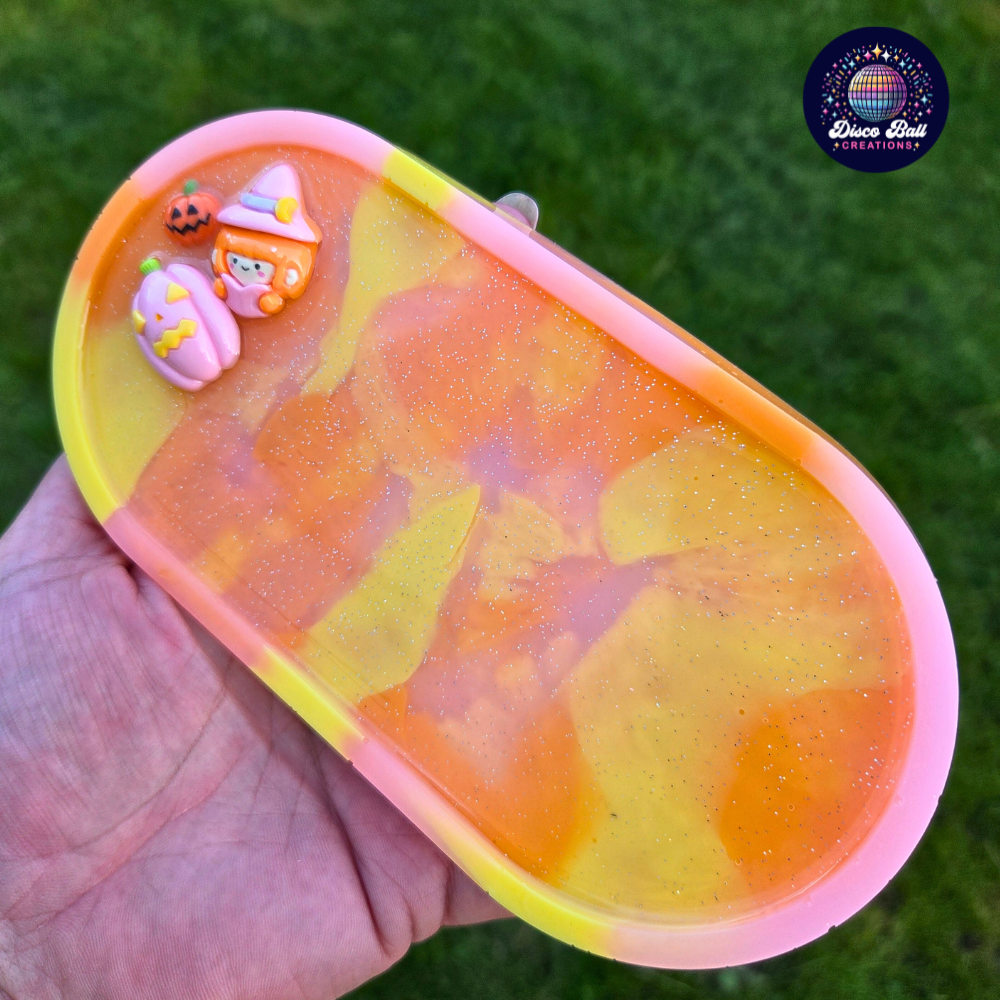 Oval trinket tray made from resin using yellow, pink and orange pastel colours with a subtle glitter in-fill and 3 character charms, a witch, a small pumpkin and a larger pumpkin