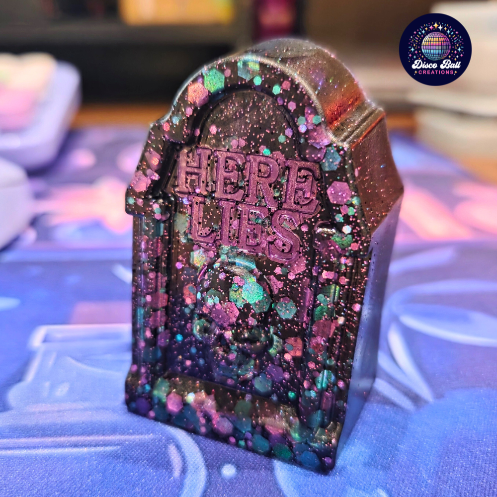 Decorative tombstone with 'Here Lies' text on a colourful glittery surface.