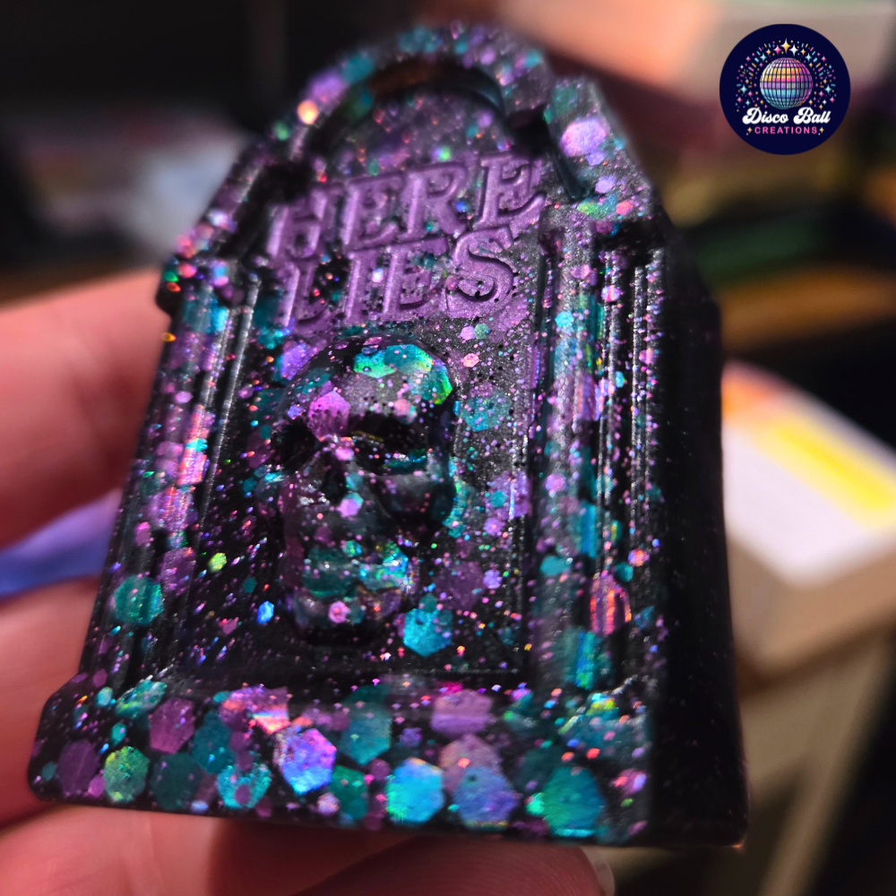 Decorative grave stone ornament with glittery finish and text 'HERE LIES', held in a hand.