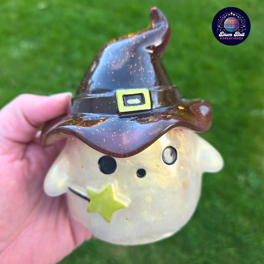 Cream Ghost figurine with Brown witch hat and gold glitter held by a hand on a grass background