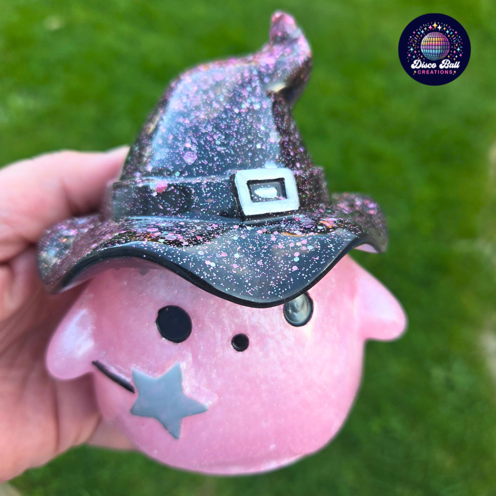 Pink character figurine with a Black glittery witch hat held against a green background