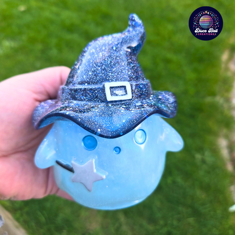 Blue glittery ghost figurine with a navy witch hat held by a hand against a green grass background.
