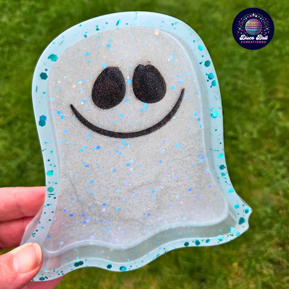 Pale blue Ghost shaped trinket tray with black eyes and smiling mouth held by a hand on a grassy background