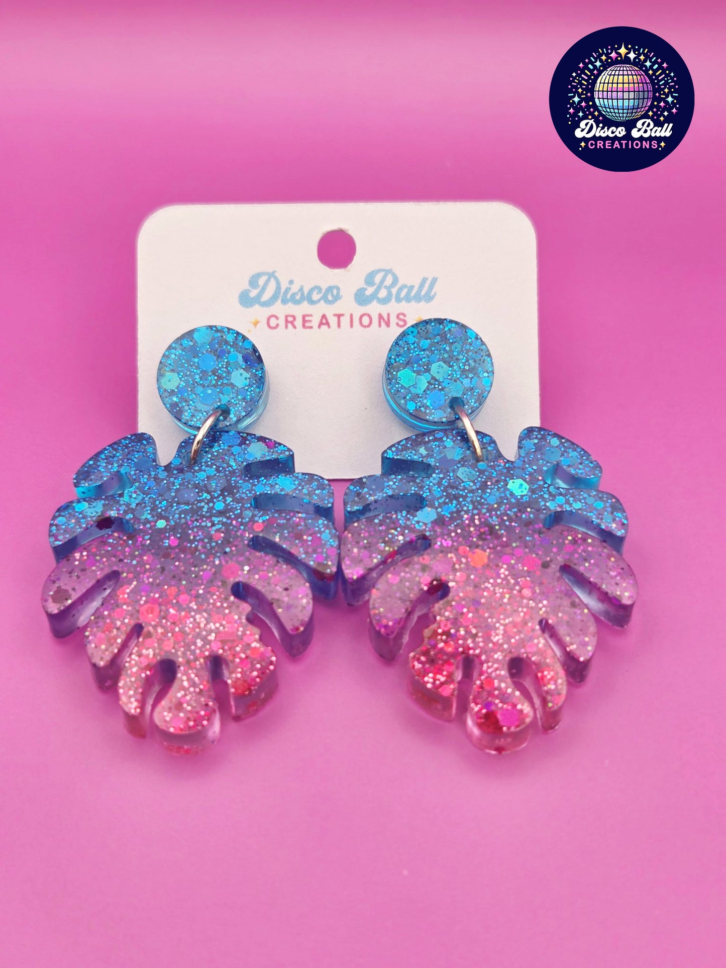 Pink and blue monstera leaf-shaped glitter resin earrings with chunky sparkle, shown on a Disco Ball Creations display card