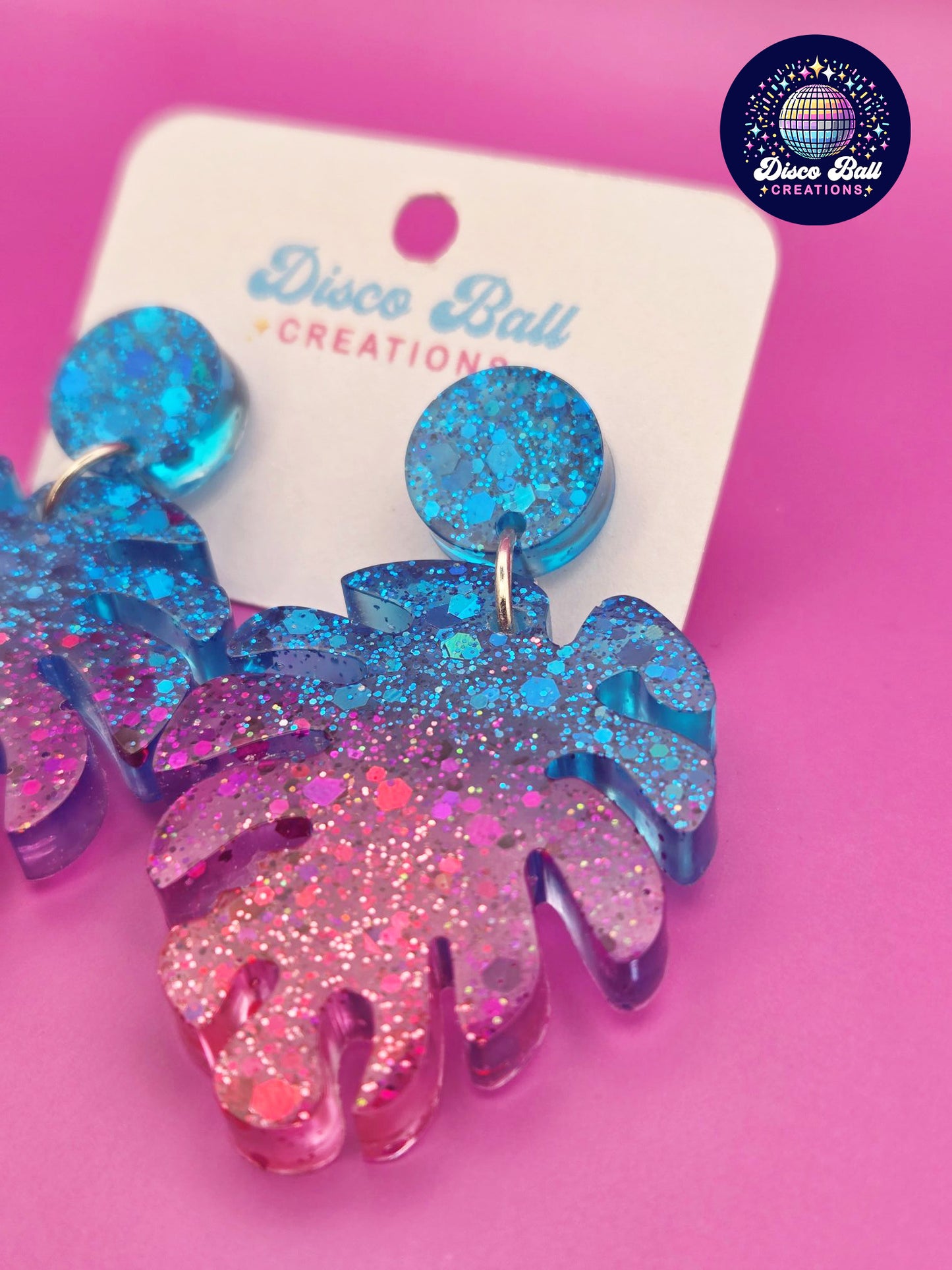 Pink and blue monstera leaf-shaped glitter resin earrings with chunky sparkle, shown on a Disco Ball Creations display card