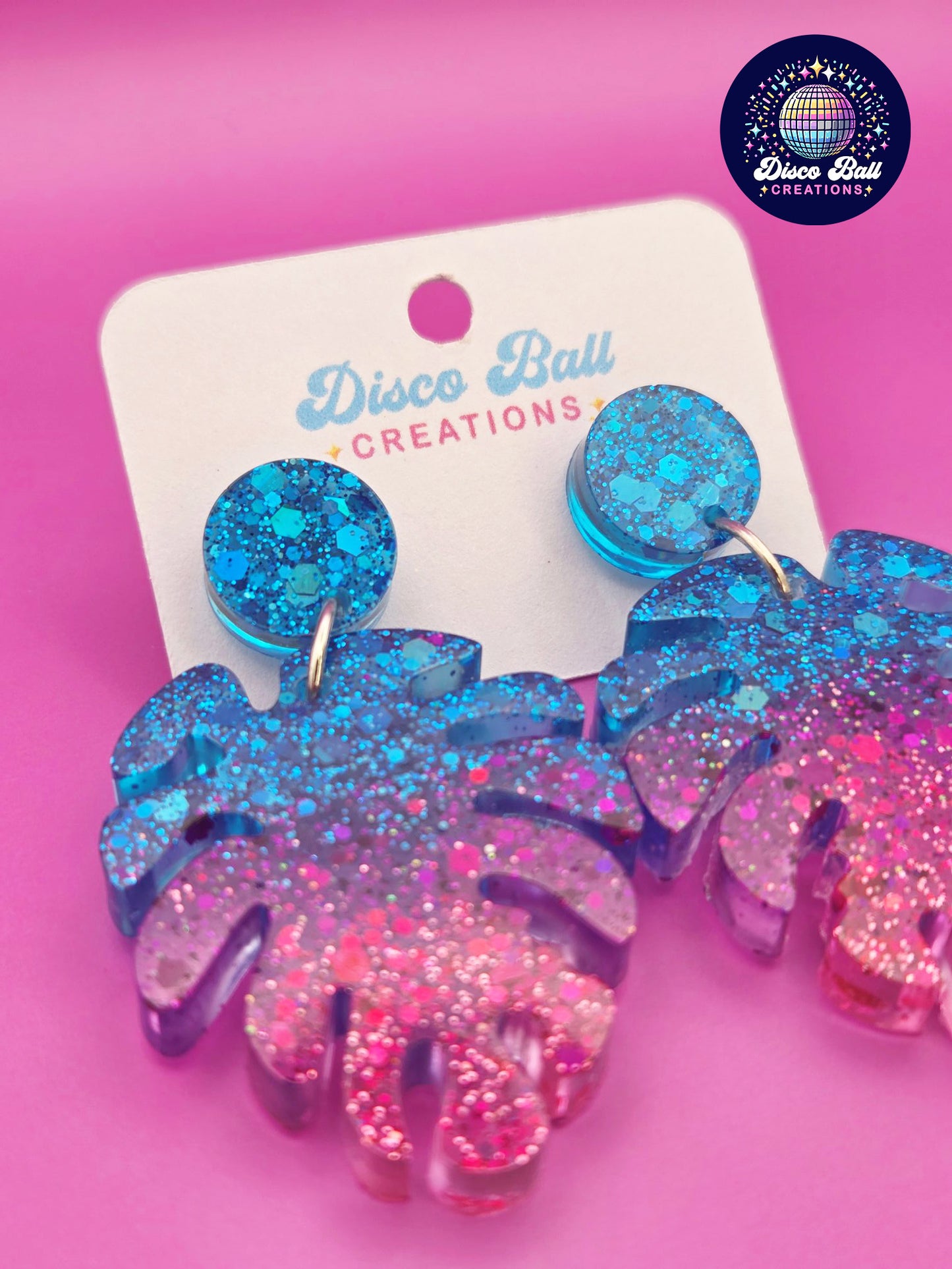 Pink and blue monstera leaf-shaped glitter resin earrings with chunky sparkle, shown on a Disco Ball Creations display card
