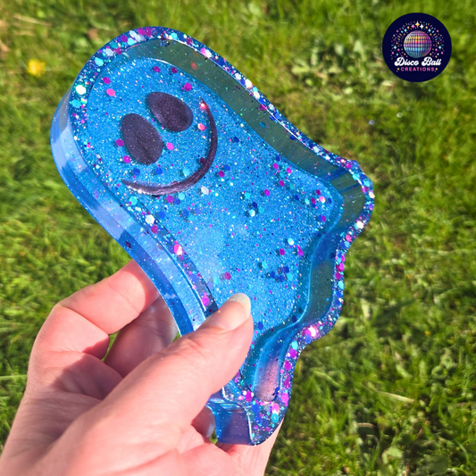 Ghost shaped blue glittery trinket tray with a smiley face held by a hand against a grass background