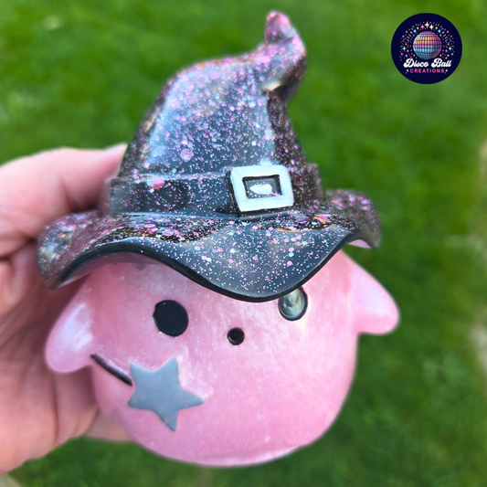 Pink character figurine with a Black glittery witch hat held against a green background