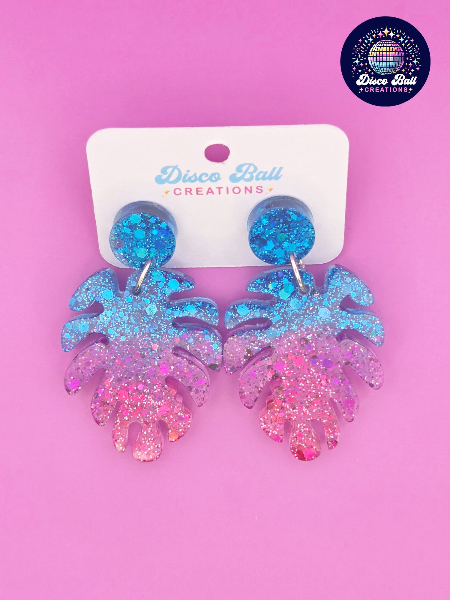 Pink and blue monstera leaf-shaped glitter resin earrings with chunky sparkle, shown on a Disco Ball Creations display card