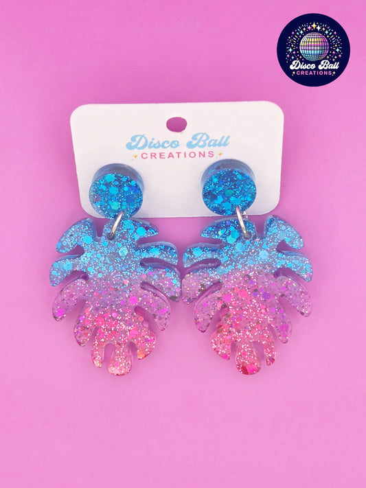 Pink and blue monstera leaf-shaped glitter resin earrings with chunky sparkle, shown on a Disco Ball Creations display card
